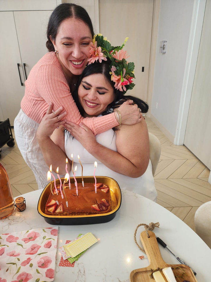 La cumpleañera, con su corona de flores, abraza a una invitada frente al pastel