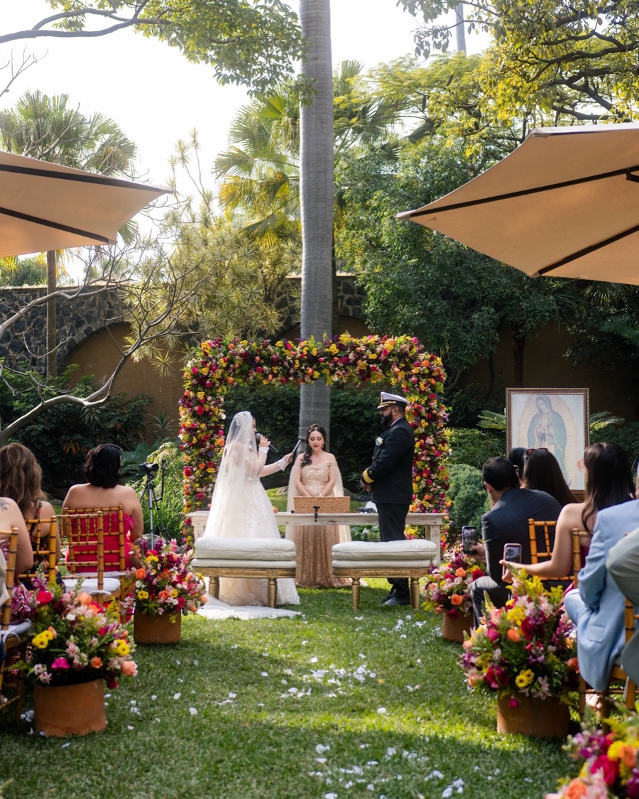 Desde el pasillo: la pareja y Sarai frente al arco de flores durante la ceremonia